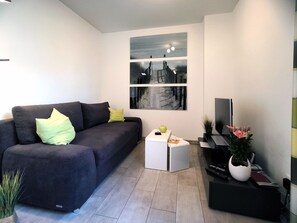 Un salon moderne avec un canapé gris, une petite table d’appoint blanche, un téléviseur à écran plat et une plante en pot.