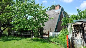 Une maison à toit de chaume, dotée d’une cheminée en briques, entourée d’une cour verdoyante et d’arbres.