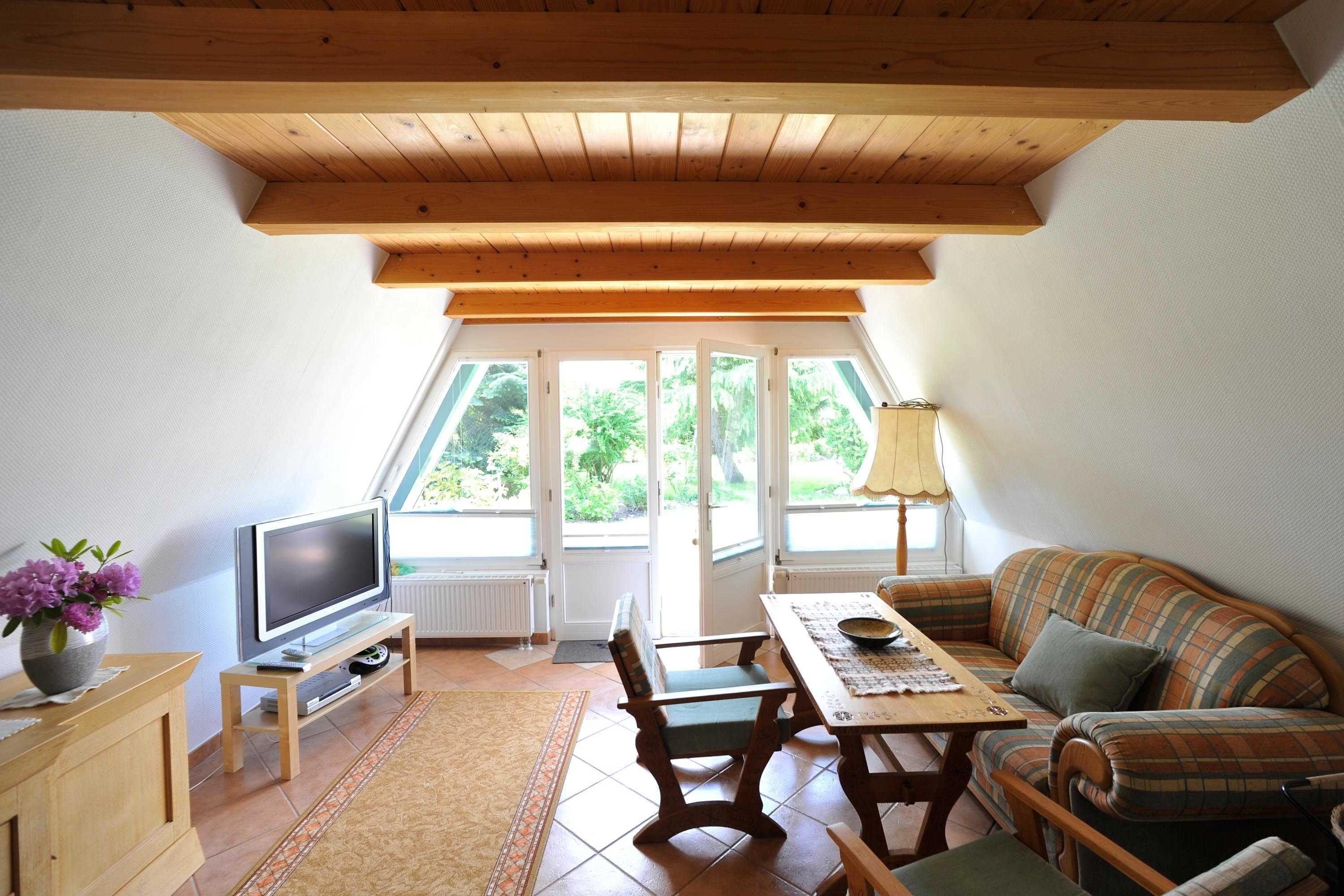 A cozy living room with a wooden ceiling, a television on a stand, a sofa, a table, and a window with a view of greenery.