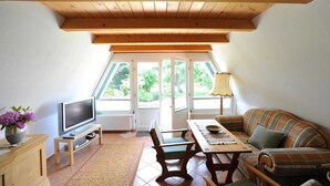 Un salon chaleureux avec un plafond en bois, un téléviseur posé sur un meuble, un canapé, une table et une fenêtre donnant sur un paysage verdoyant.