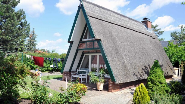 Une maison à toit de chaume, dotée d’une cheminée en briques, entourée d’un jardin où l’on trouve des plantes en pot et un fil à linge.