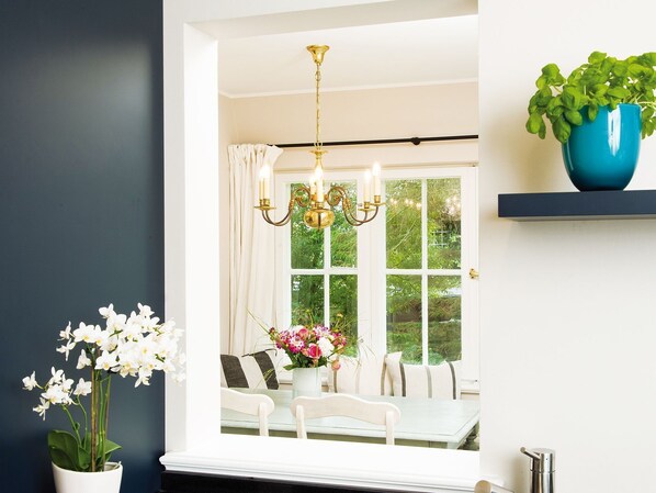 A dining area with a white table and chairs, a chandelier, a potted plant, and a window with curtains.