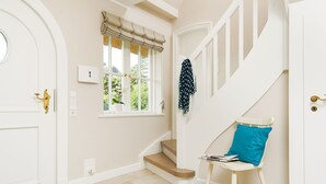 Ein Flur mit einer weißen Tür, einem Fenster mit Vorhang, einer Treppe und einem Sessel mit Kissen.