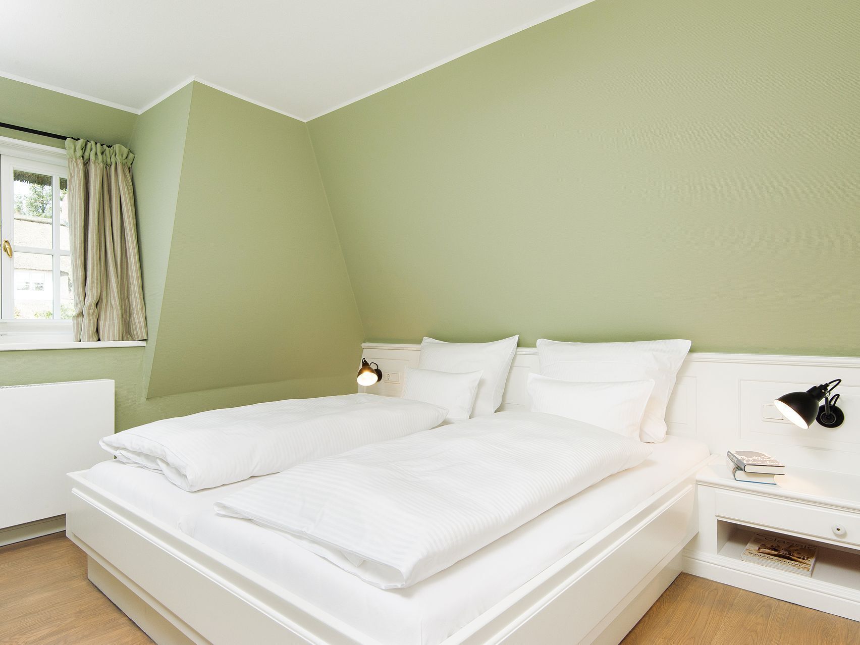 Ein Doppelbett mit weißer Bettwäsche in einem Zimmer mit grünen Wänden und einem fensterbelaubten Fenster.