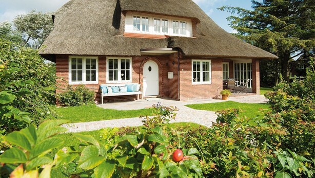 Ein traditionelles Backsteinhaus mit einem Strohdach, einer Veranda mit einer Bank und einem gepflegten Garten.