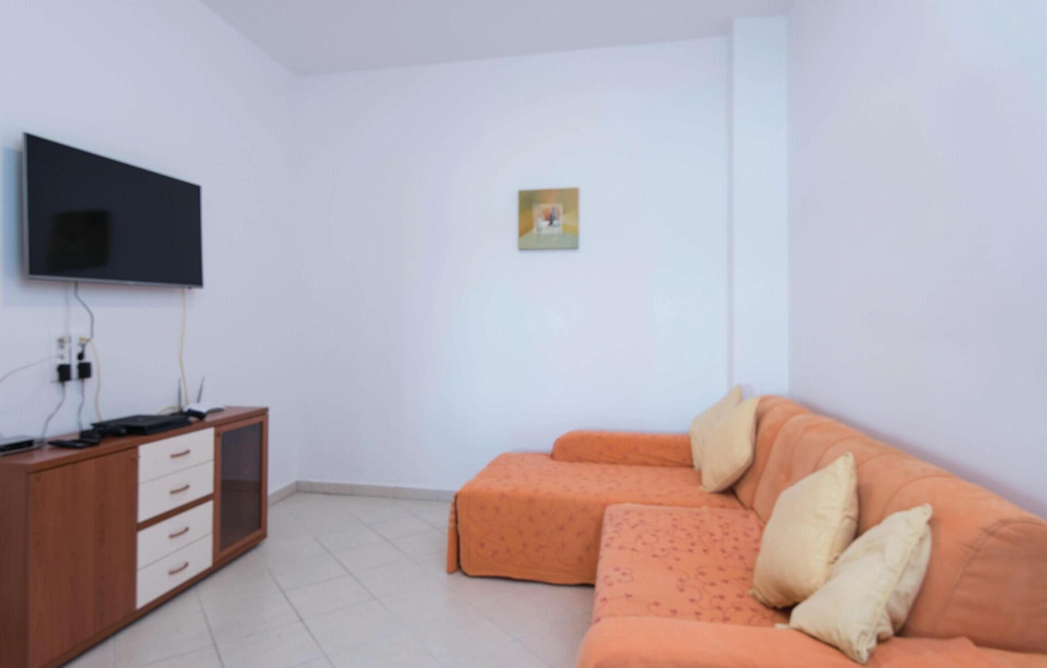 Habitación con una televisión de pantalla plana montada en la pared, un mueble de madera con cajones, un sofá naranja con cojines y una pequeña foto enmarcada en la pared.