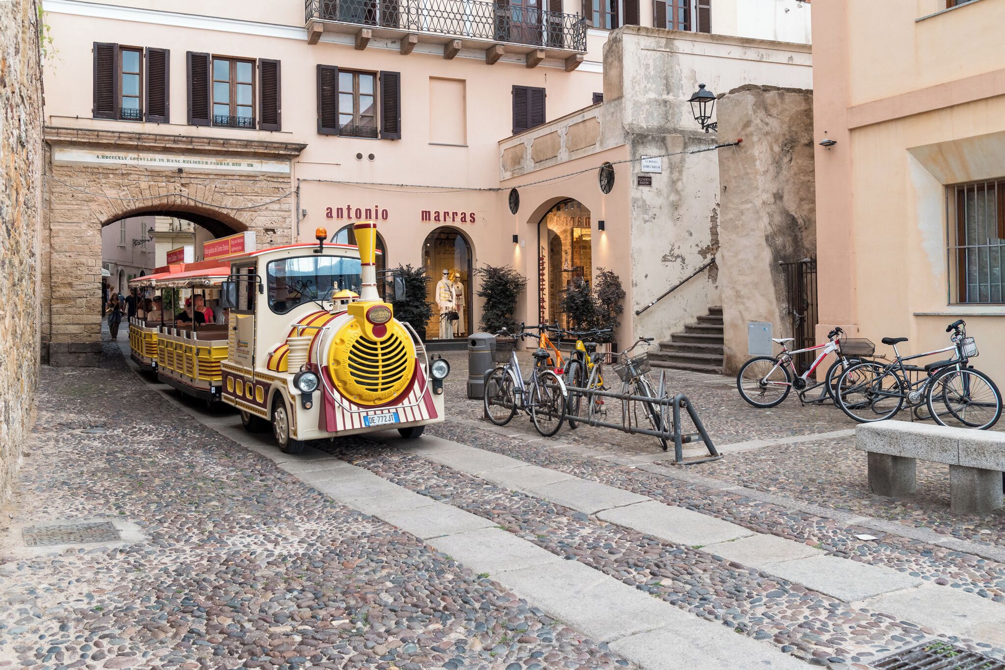 A vintage train in a narrow street, a building with 'antonio marras' signage, and bicycles parked nearby.