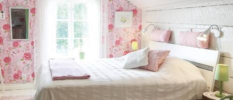A bedroom with a sloped ceiling, floral wallpaper, a bed with white bedding, a green plant, and a window with a view of greenery.