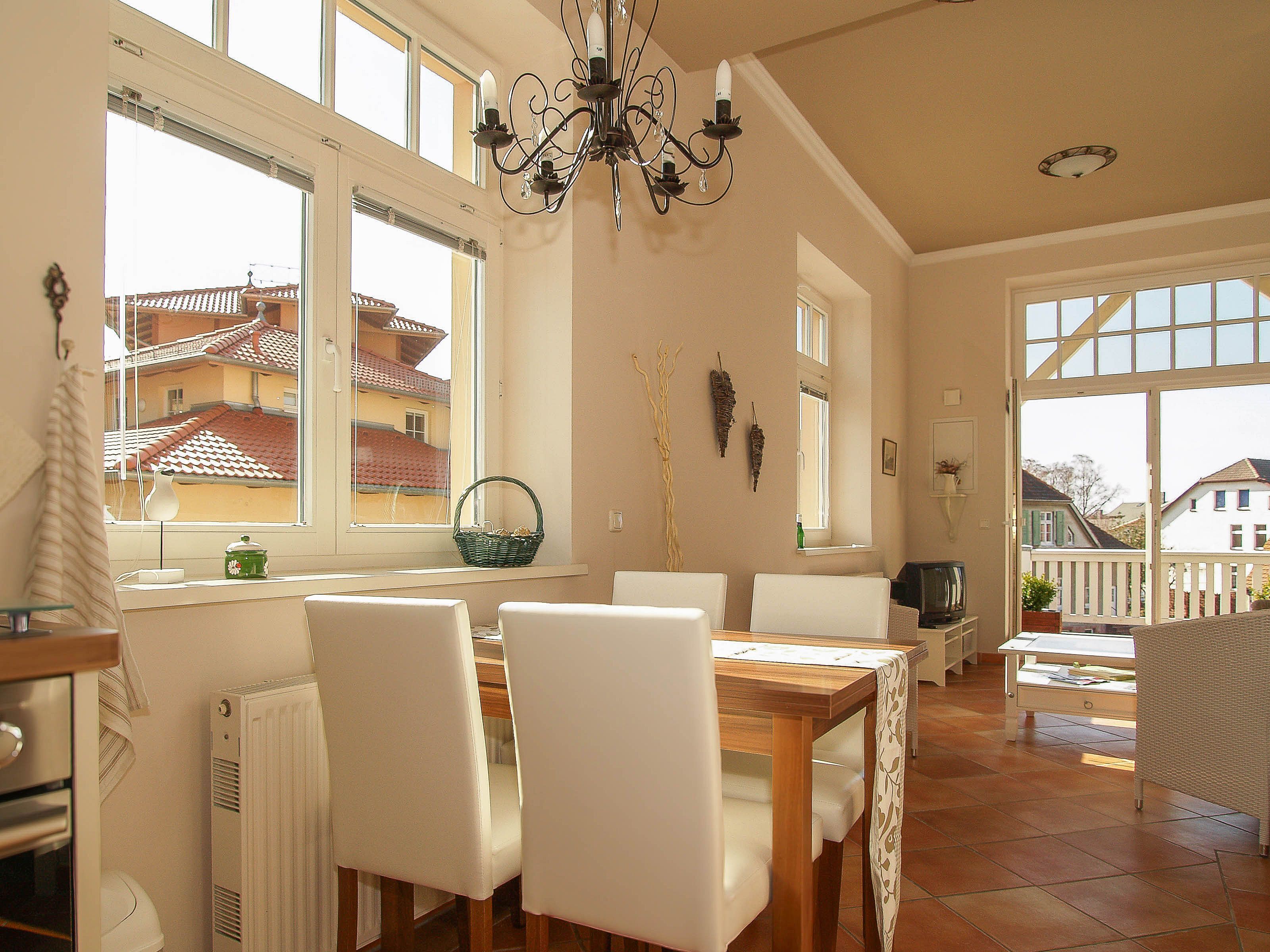 Une salle à manger avec une table en bois et des chaises blanches, de grandes fenêtres donnant sur des maisons et un lustre.