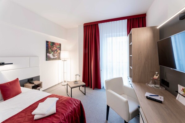 A hotel room with a bed, a desk, a chair, a TV, and a window with red curtains.