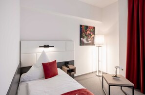 A hotel room with a bed, a bedside table, a lamp, a painting, and a red curtain.
