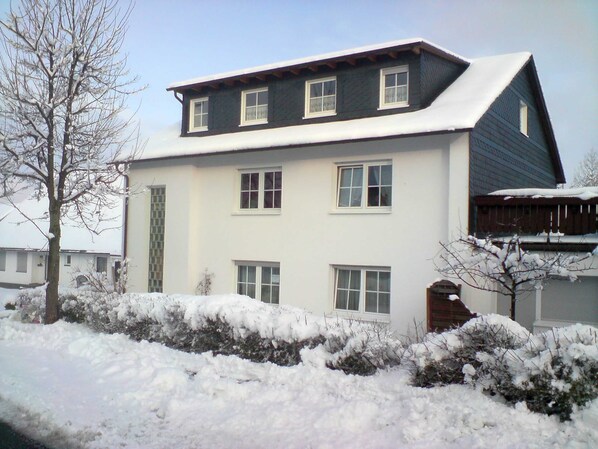 A two-story house with a white exterior, multiple windows, and a sloped roof covered in snow.