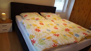 Ein Bett mit floraler Tagesdecke, ein Nachttisch mit Lampe und ein Fenster mit Vorhängen.