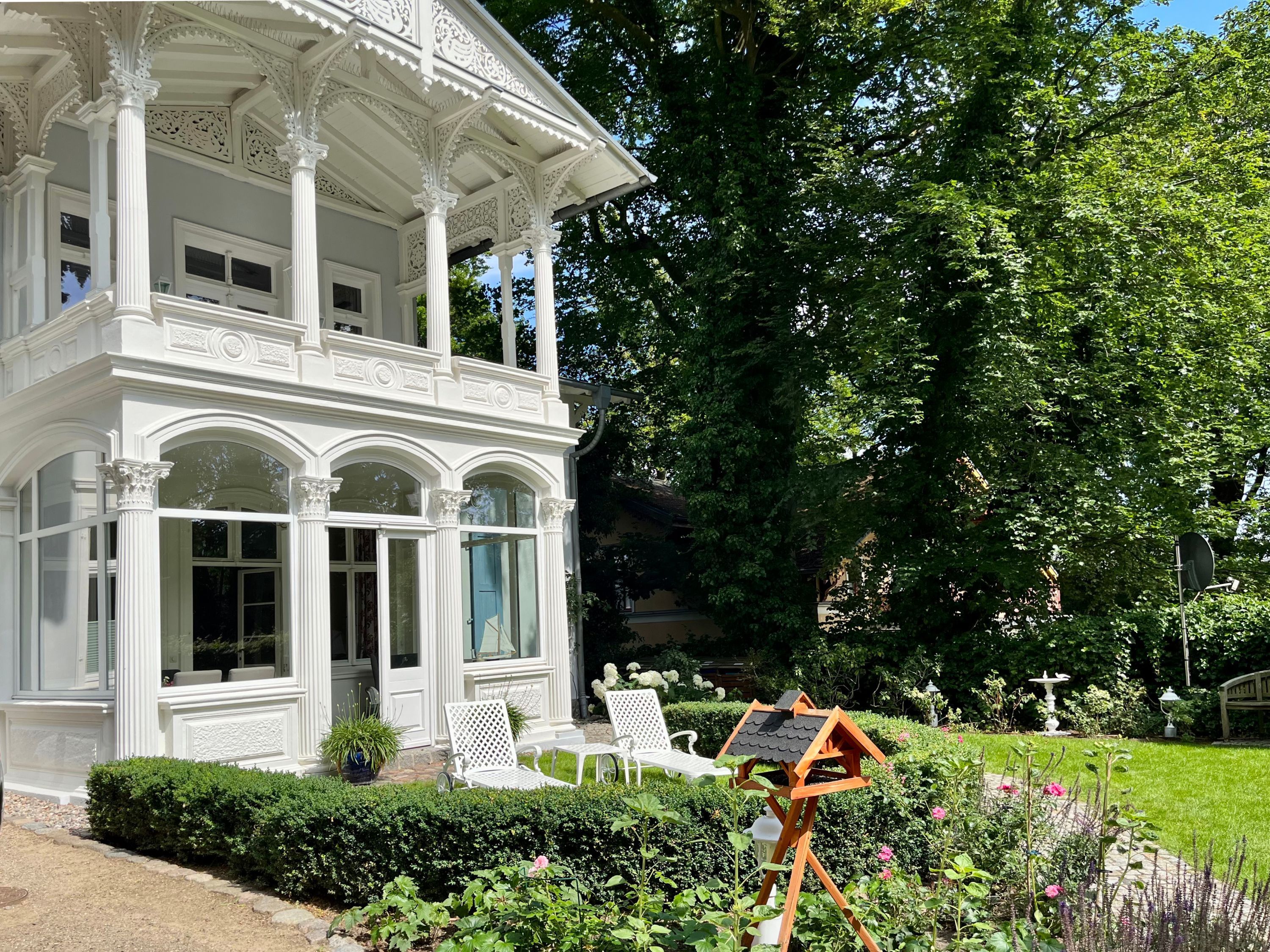 Un belvédère blanc, orné de colonnes décoratives et doté d’un porche abrité, est entouré d’un jardin bien entretenu, agrémenté de haies et de plantes à fleurs.
