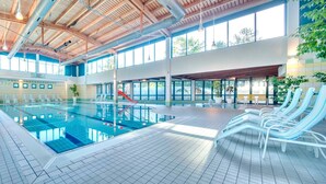 En inomhuspool med kristallklart blått vatten, omgiven av vita solstolar och krukväxter.