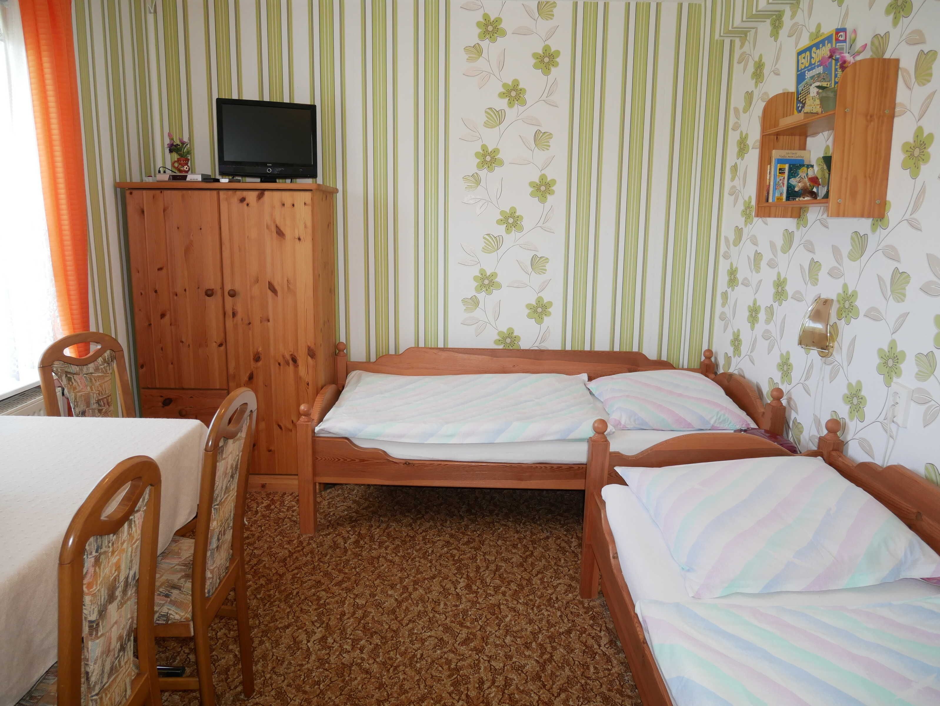 A bedroom with a wooden bed, a TV on a wooden cabinet, a small shelf with books, and a striped wallpaper.