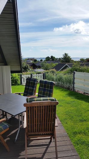 Eine Terrasse mit Tisch und zwei Stühlen, ein weißer Zaun und Blick auf Häuser sowie ein Gewässer.