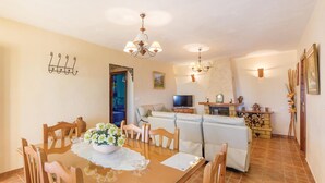 A dining area with a wooden table, chairs, and a chandelier. There is a sofa and a television in the background. - Amazing home in Corumbela (Corumbela)