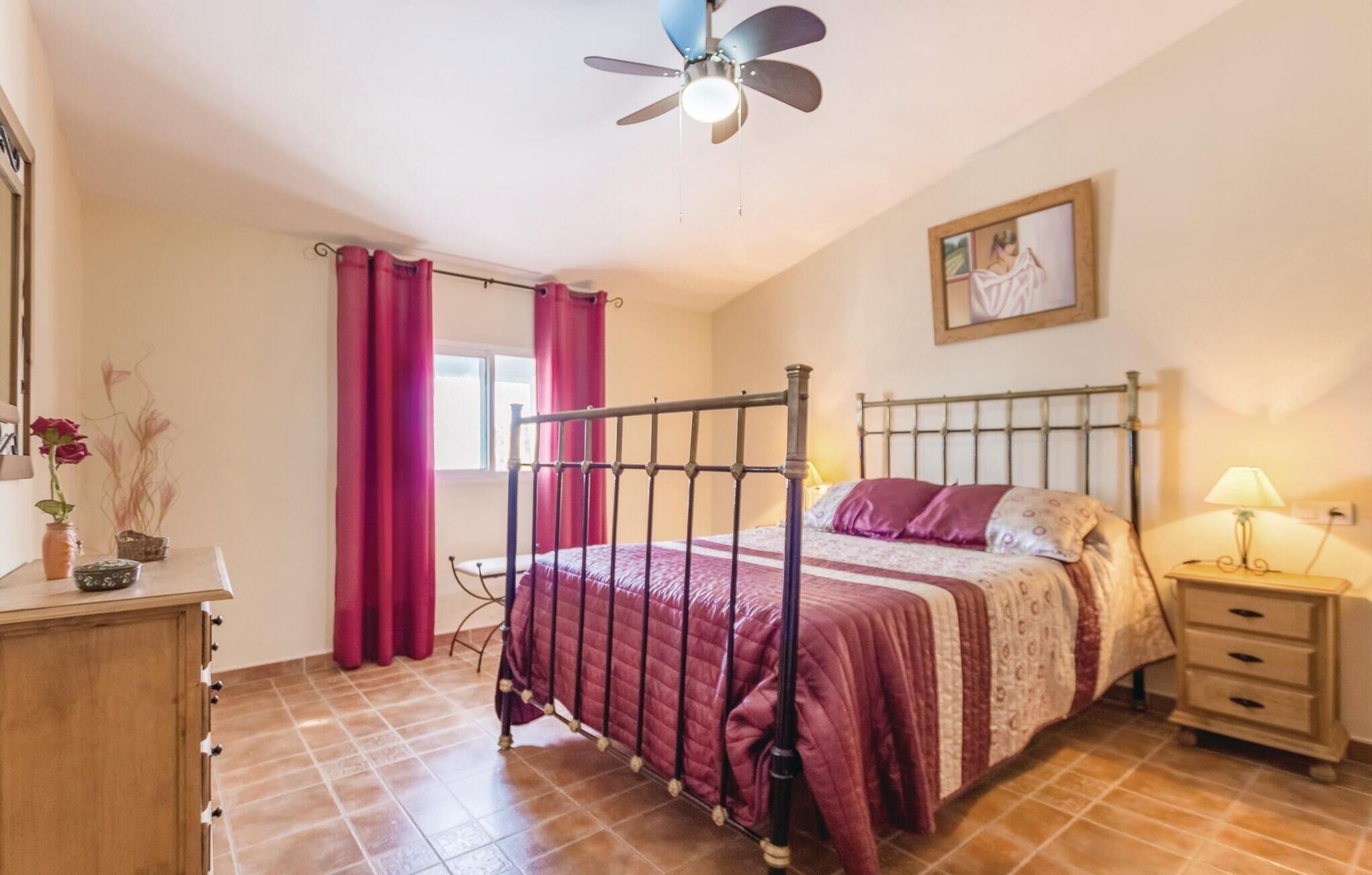 Une chambre à coucher comprenant un lit, une table de chevet, une commode, un ventilateur de plafond et une fenêtre avec des rideaux.