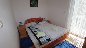 A single bed with a patterned bedspread, two bedside tables with lamps, a small bedside table with a lamp, and a window with sheer curtains.
