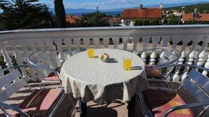 Ein runder Tisch mit zwei Stühlen, einer Tischdecke und zwei Gläsern Orangensaft auf einem Balkon mit weißem Geländer.