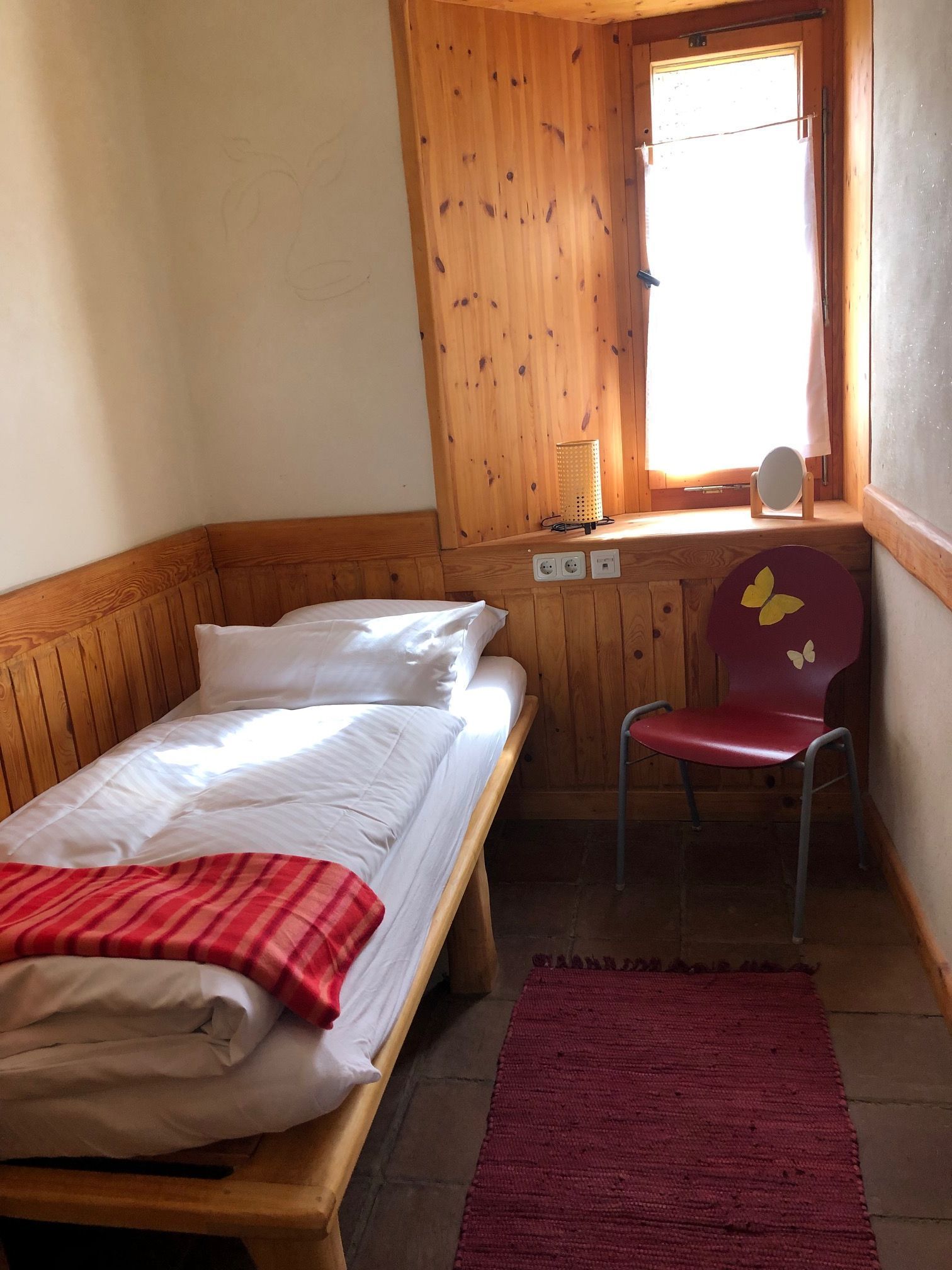 A single bed with a red blanket, a wooden chair with a butterfly design, a window with a curtain, and a wall-mounted lamp.