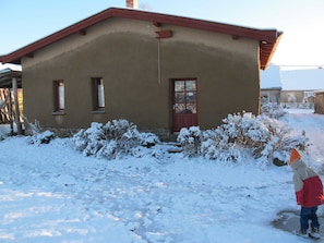 A single-story building with a red door and windows, surrounded by snow-covered ground and bushes.