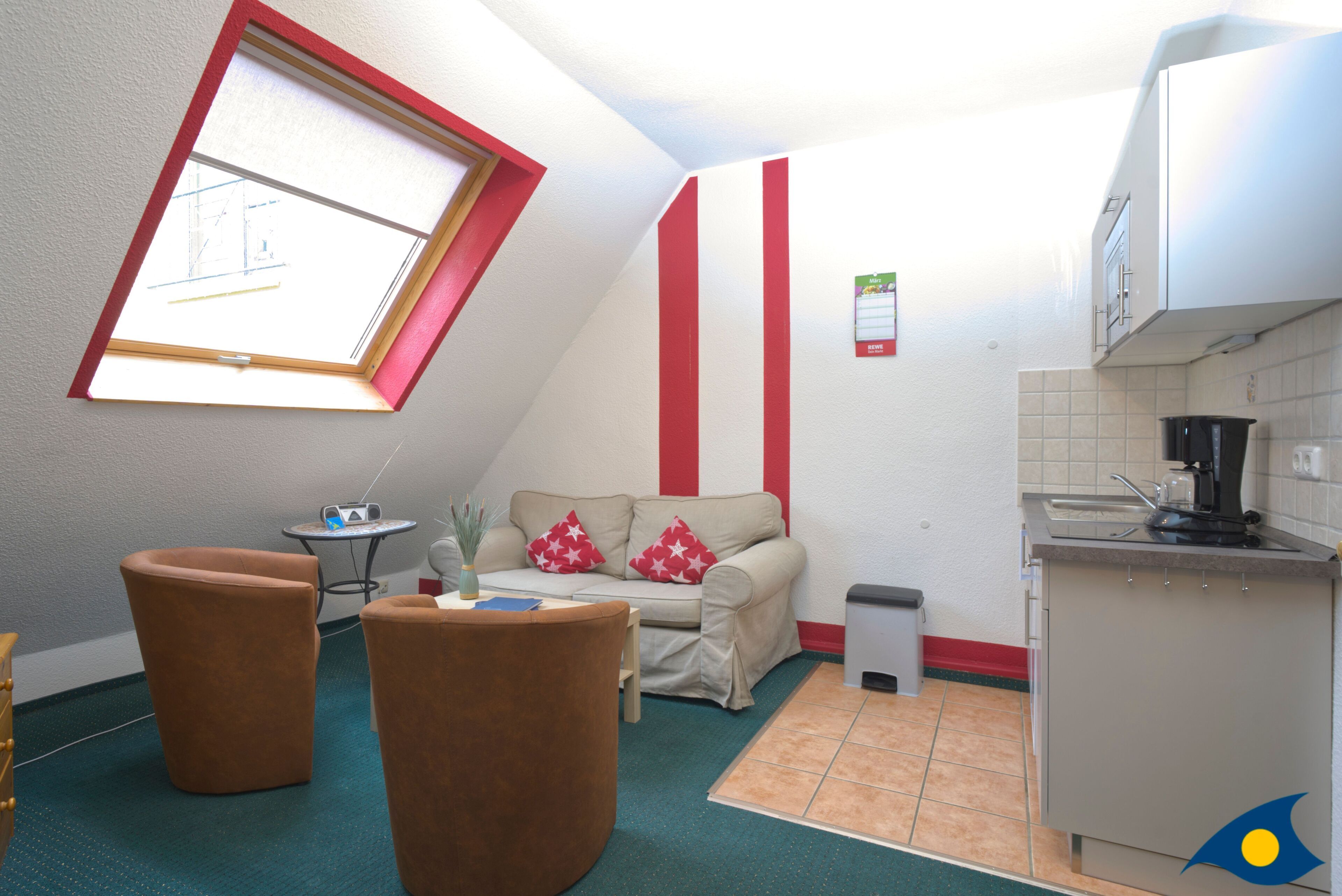 Un espace de vie compact comprenant un coin cuisine, un canapé avec des coussins rouges et blancs, une petite table et une fenêtre avec un cadre rouge.