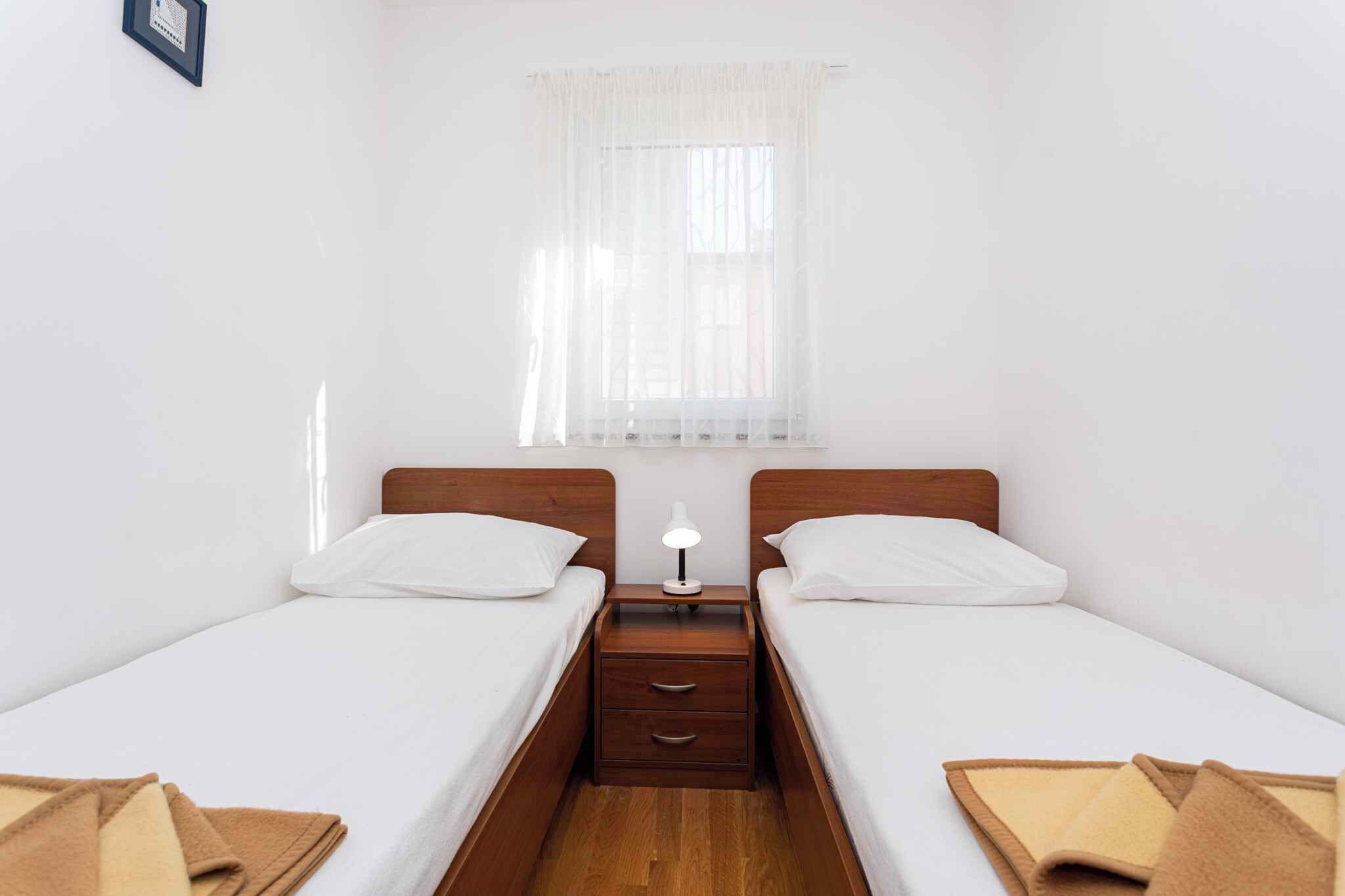 Une petite chambre propre, avec deux lits simples, une table de chevet avec une lampe et une fenêtre ornée de rideaux légers.