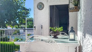 Ein weißer Outdoor-Tisch mit Glasplatte, ein weißer Metallstuhl, ein Blumentopf, ein Laternenlicht und eine dekorative Wandplatte.