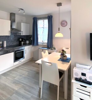 A modern kitchen with a dining area, a clock, and a window with curtains. - Haus Meeresblick FeWo Seeschwalbe A 2.21 Ref. 128704 - FeWo Seeschwalbe A 2.21 (Baabe)