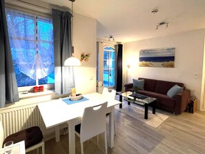 A living room with a white table, a brown sofa, a coffee table, and a window with curtains. - Haus Meeresblick FeWo Seeschwalbe A 2.21 Ref. 128704 - FeWo Seeschwalbe A 2.21 (Baabe)