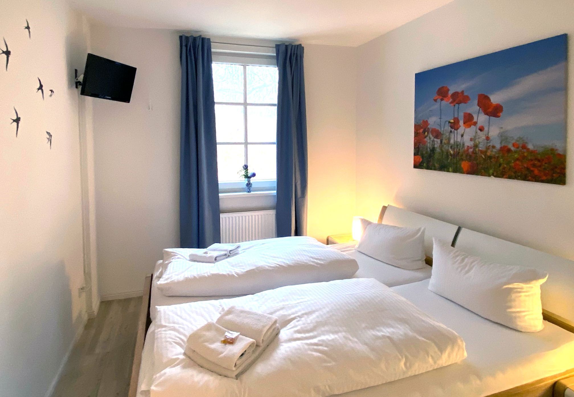 Habitación de hotel con dos camas, un televisor de pantalla plana montado en la pared, una ventana con cortinas azules y un cuadro de amapolas en la pared.