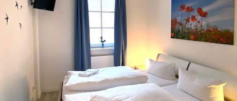 A hotel room with two beds, a flat-screen TV mounted on the wall, a window with blue curtains, and a painting of poppies on the wall.