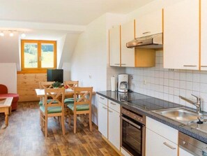 Una cuina compacta amb zona de dinar, forn de pared i finestra amb vista a vegetació.
