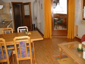 Un comedor con mesa y sillas de madera, una cocina al fondo y vistas a un balcón con una silla a través de un ventanal.