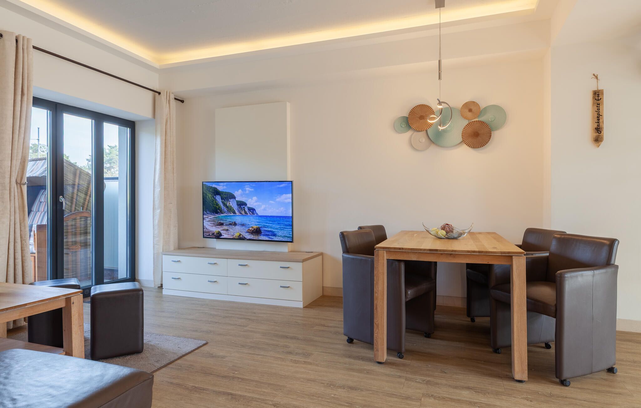 Un salon moderne doté d’un téléviseur à écran plat, d’une table à manger en bois, de fauteuils en cuir, d’un meuble blanc et d’un balcon agrémenté de rideaux.