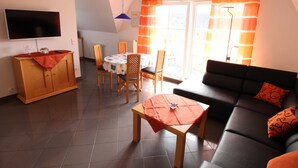 Ein Wohnzimmer mit einer Couch, einem Esstisch mit Stühlen, einem an der Wand montierten Fernseher und einem Fenster mit Vorhängen.