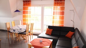 Ein Wohnzimmer mit einer schwarzen Couch, orangefarbenen Kissen, einem Holztisch und einem Esstisch mit Stühlen.