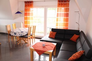 Una sala de estar con un sofá negro, cojines naranjas, una mesa de centro de madera y una mesa de comedor con sillas.