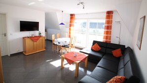 Ein Wohnzimmer mit einer schwarzen Couch, einem Holztisch und einem an der Wand befestigten Fernseher.