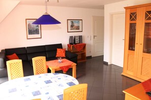 Una sala de estar con un sofá negro, una mesa redonda con mantel estampado, un aparador de madera y dos cuadros enmarcados en la pared.