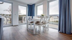 A modern dining area with a white table and chairs, large windows with blue curtains, and a view of the outside. - Villa Margarete Whg. 8 - Villa Margarete, BEACH NEXT (Heringsdorf)
