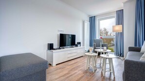 A modern living room with a flat-screen TV, a white entertainment unit, a grey sofa, a grey armchair, and a small table with a lamp. - Villa Margarete Whg. 8 - Villa Margarete, BEACH NEXT (Heringsdorf)
