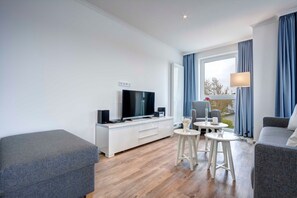 Ein modernes Wohnzimmer mit Flachbildfernseher, einer weißen Unterhaltungsecke, einem grauen Sofa, einem grauen Sessel und einem kleinen Tisch mit einer Lampe.