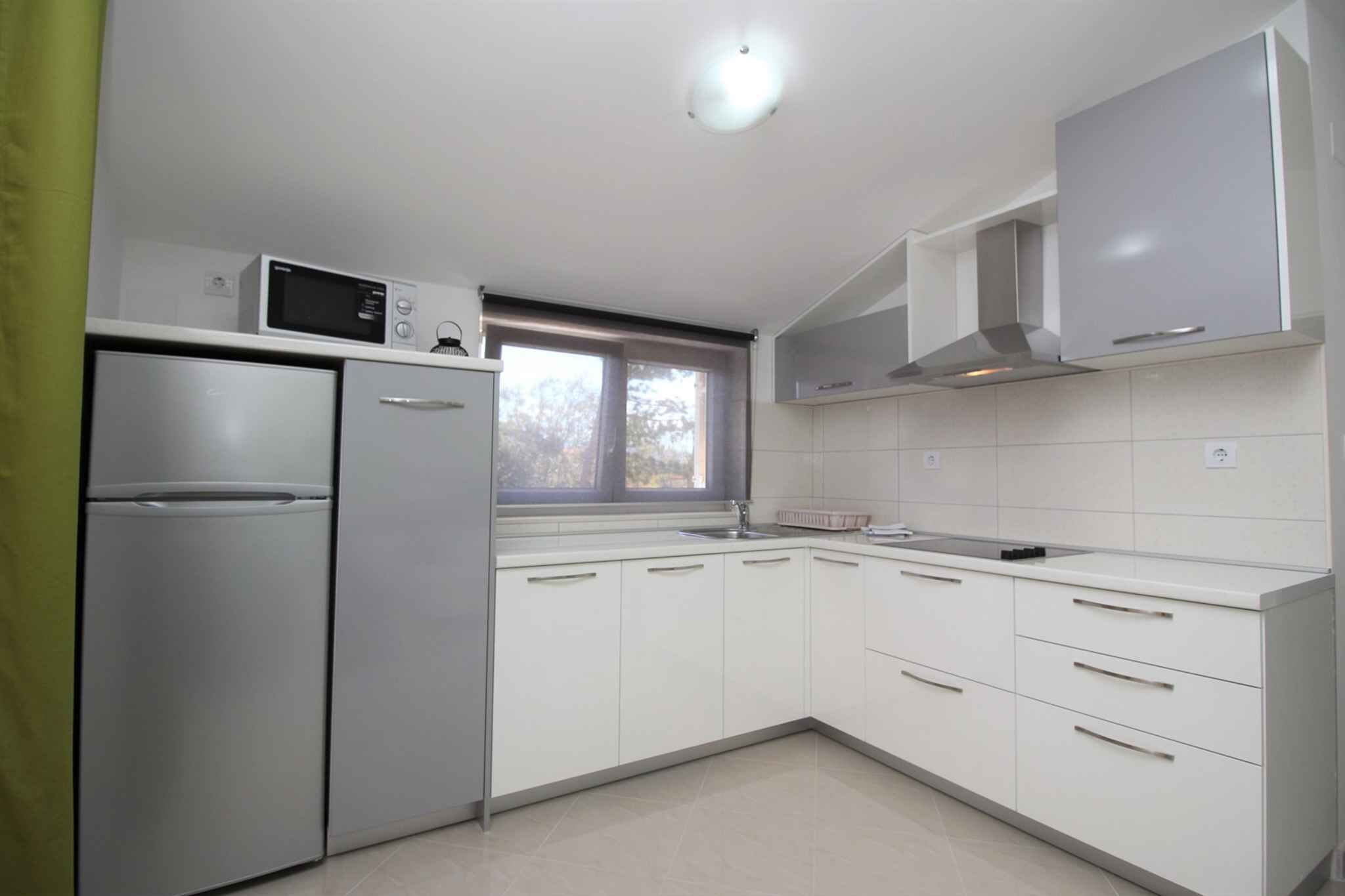A modern kitchen with white cabinets, a stainless steel refrigerator, a microwave, and a window with a view of trees.