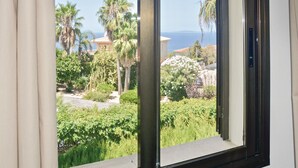 A view through a window with a view of a garden, palm trees, and a building with a red roof. - Stunning home in Bahia Azul-Badia Blava (Bahia Azul-Badia Blava)