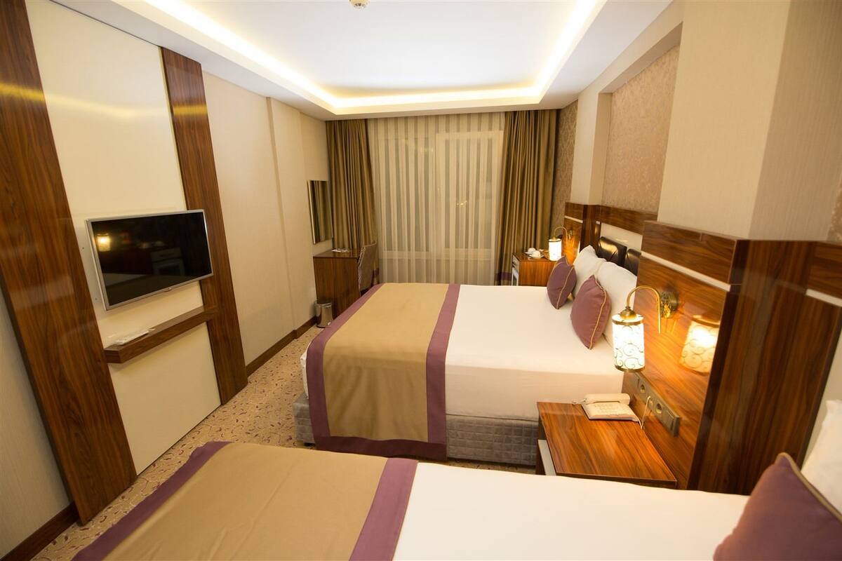 A hotel room with two beds, a TV, a bedside table with a lamp, and a window with curtains.