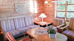 En mysig stuga med en randig soffa, ett träbord med en metallskål och blommor, och ett fönster med utsikt över grönskan.
