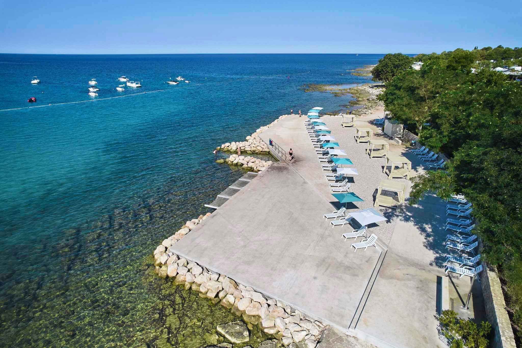 An aerial view of a beach with clear blue water, a concrete walkway, and rows of sun loungers.